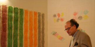 Man with glasses browsing a book. In front of him a drawing hangs on the wall with red, green, orange and brown strips