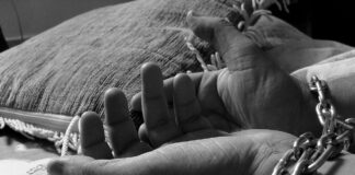 The picture is a black and white photography showing a pair of chained hands near a pillow on a bed cover with infantile decorations