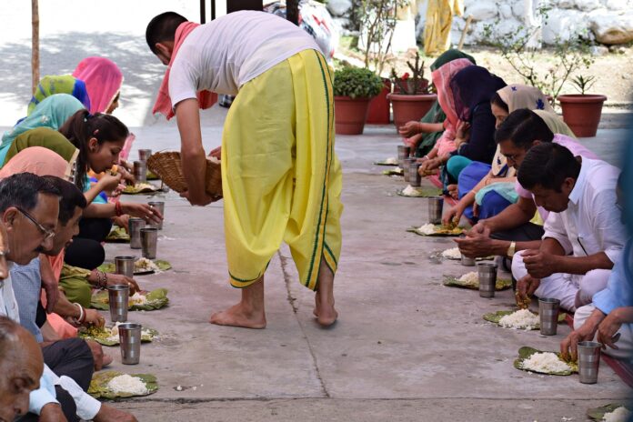 The photo represents an Indian man dressed in white and yellow providing food to a couple of parallel lines of people. The meal consist of a dish with some rice and a iron glass of water